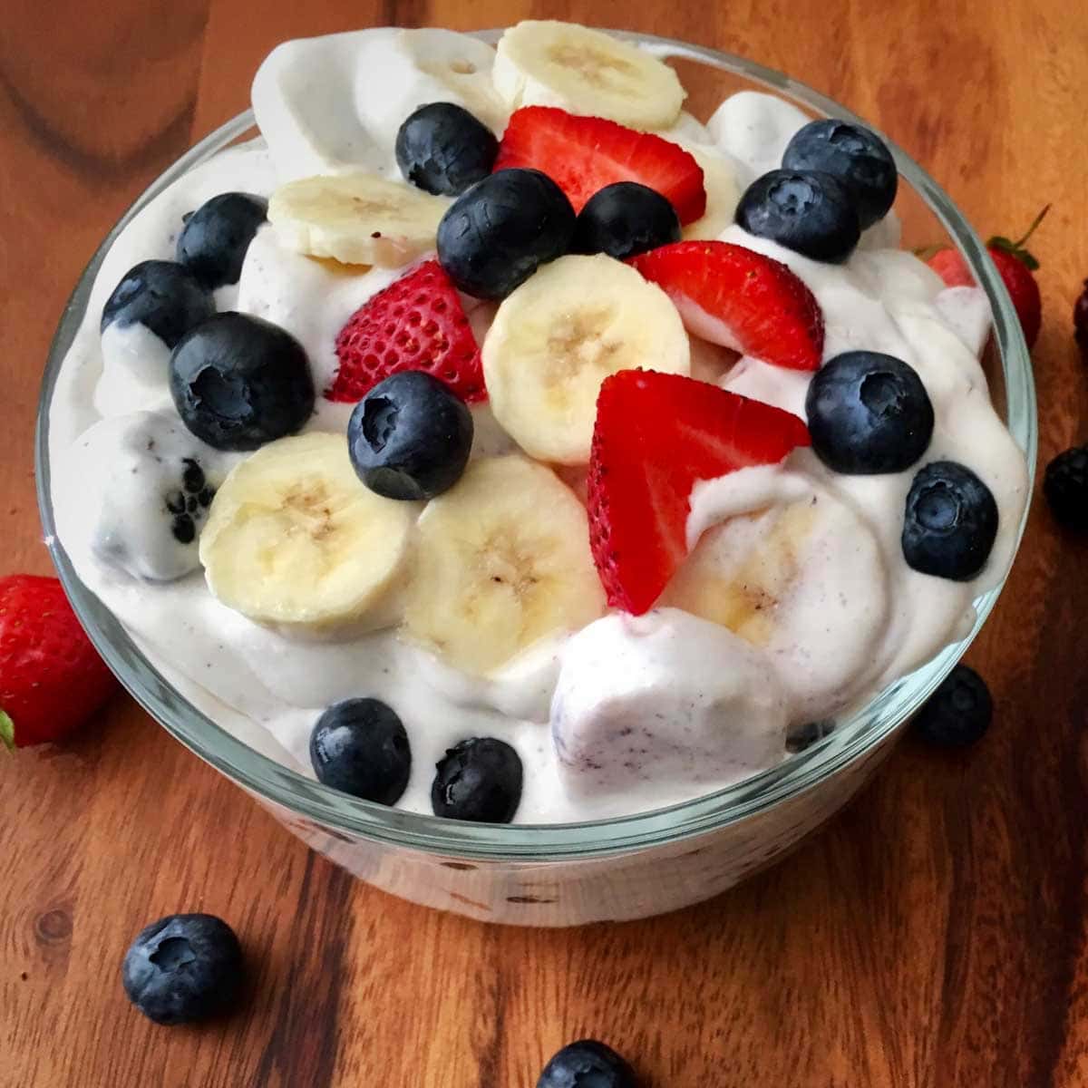 cheesecake fruit salad in a  bowl garnished with fruit on a top and surrounded by blueberries on a wood cutting board