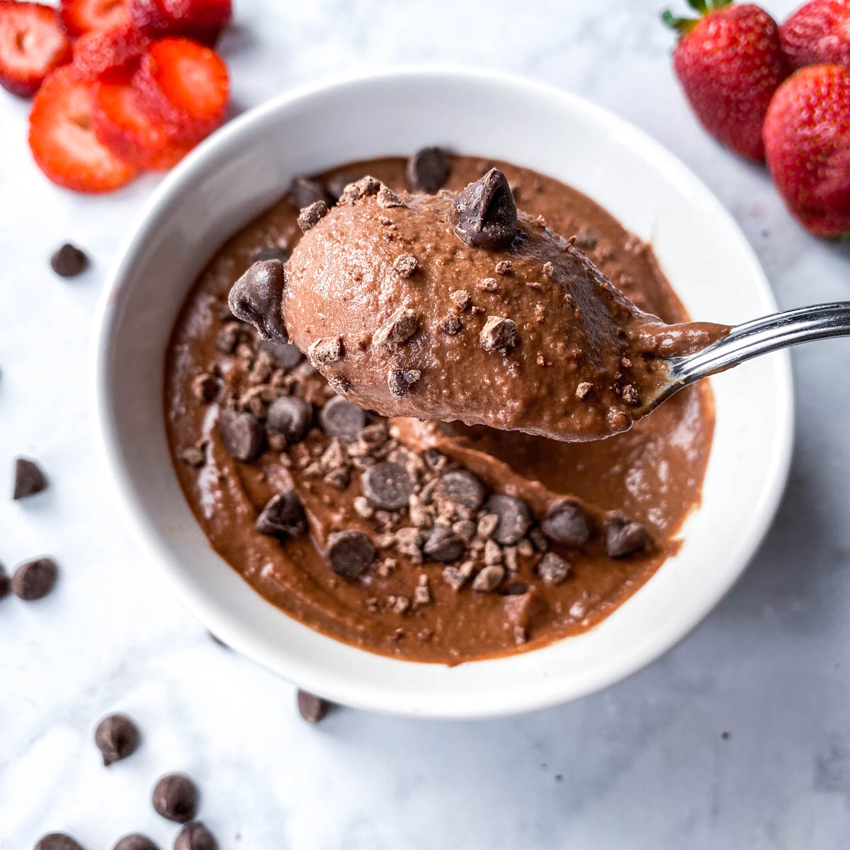 brownie batter hummus with a spoon dipping it out of a bowl. Strawberries in the background. 